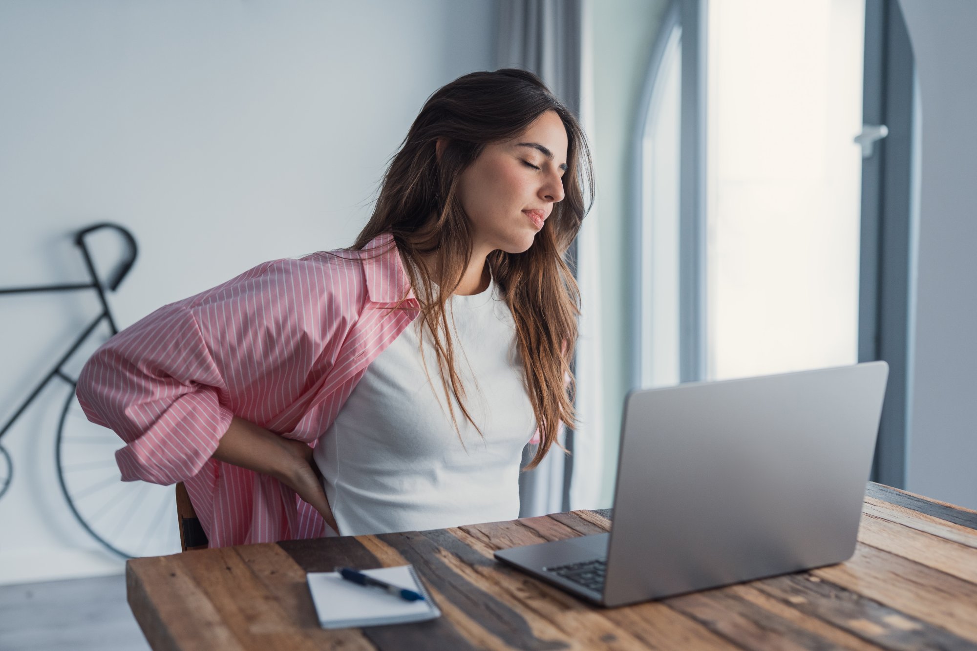 Woman at her desk holding her lower back in discomfort, a common reason patients explore Emsculpt NEO for back pain in Katonah, NY